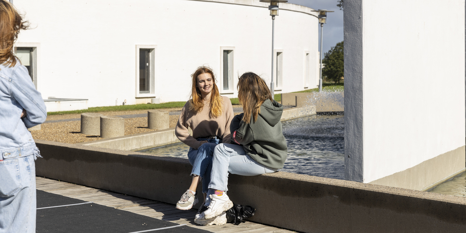To studerende sidder udenfor ved et springvand. En tredje studerende står i udkanten af billedet. Jeans, beige sweater, grøn hættetrøje, rødt label, hvide sneakers, træer, lygtepæle, sort taske. 