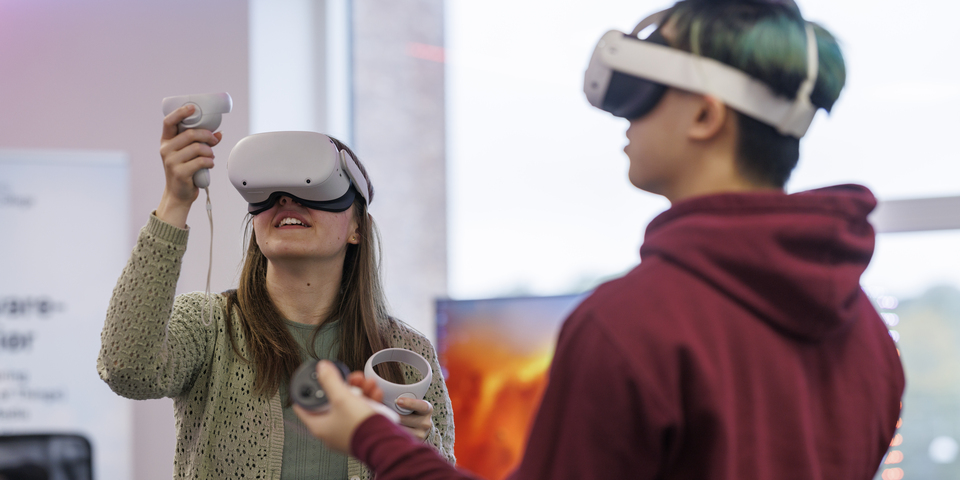 En kvindelig og en mandlig studerende står med hvide VR-briller på og fjernbetjeninger i et undervisningslokale. Lamper, neonlys, LED-lyskæde, fjernbetjening, remote, teknologi, digital, rød hættetrøje, gul cardigan, tv-skærm, jeans. 