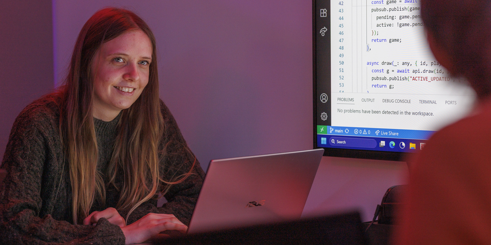 En kvindelig studerende sidder ved et bord med computere foran en stor computerskærm, der viser koder, kodningsprogram i et lokale oplyst af rødt og blåt lys. Gaming, computer, bærbar, briller, hjul, jeans, orange sweater. 