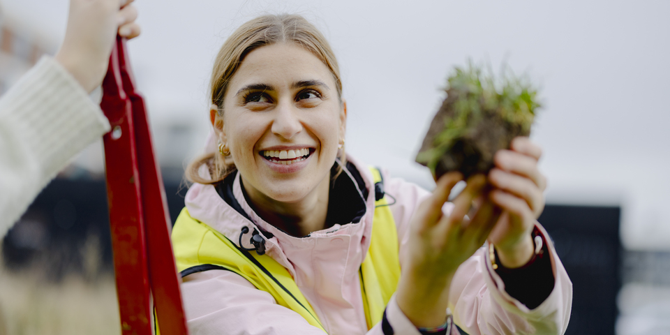 To kvindelige studerende har refleksveste på og har gravet i jorden for at kunne foretage målinger med måleudstyr med ledning. Jeans, lyserød jakke, sneakers, bygninger, nybyggeri, ledning, prøve, arbejde, praksis, spade, tang, græs, sort spand, briller, rød kabelrulle, parkeringsplads, jordprøve. 