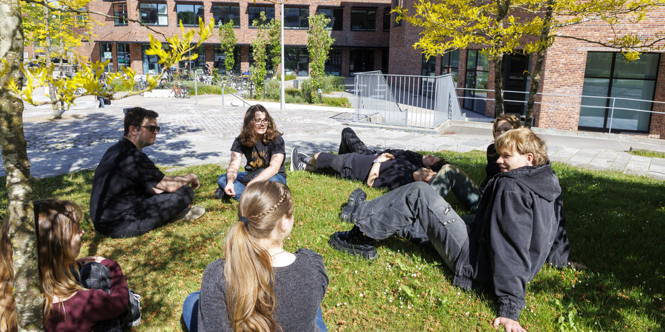 En gruppe af studerende sidder på græsplænen udenfor. Træer, bygninger, campus Horsens, jeans, fritid, forår, pause, fritid, internationale studerende. 